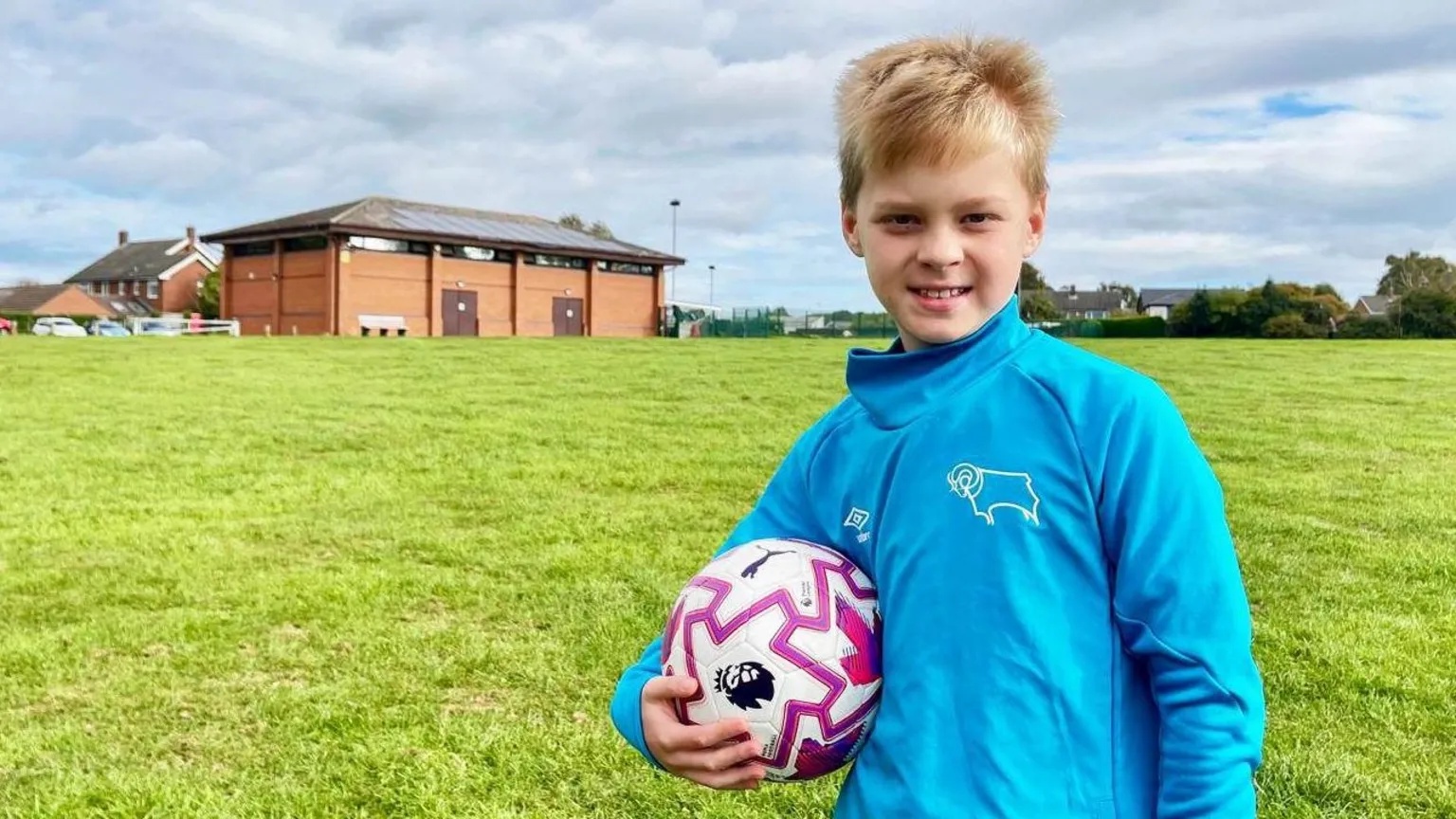 A boy in a blue tracksuit top is holding a football on a grassy field with a sports centre and floodlights in the background.