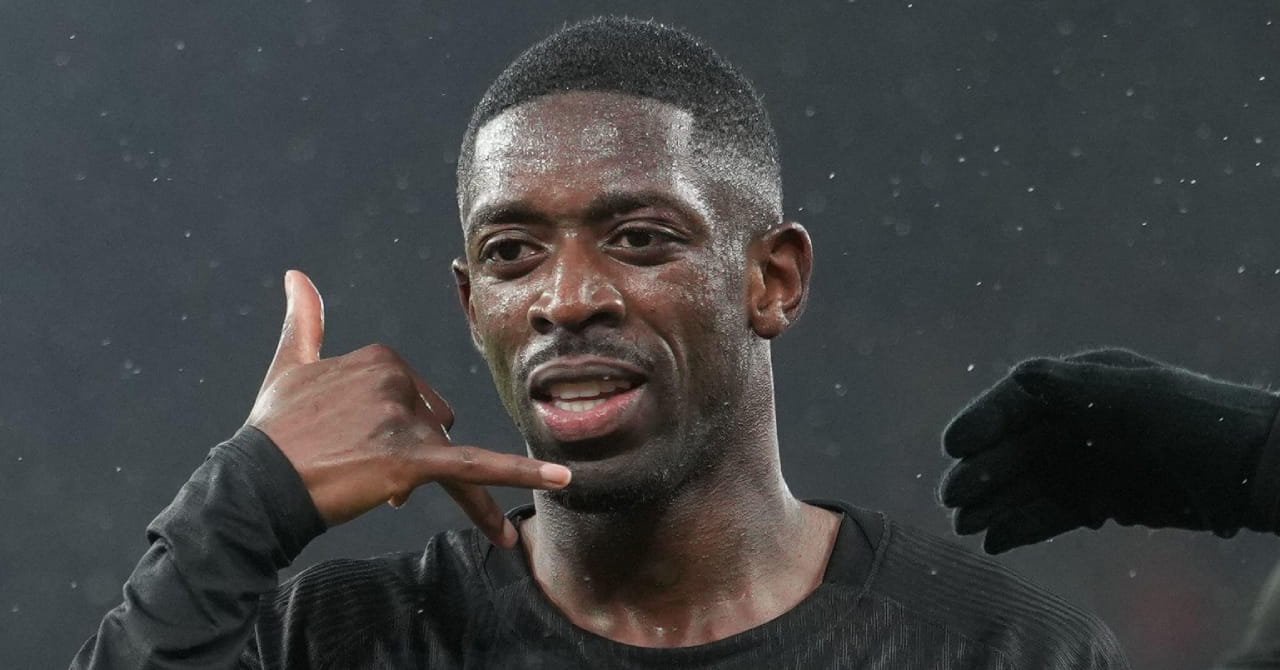 Paris Saint-Germain footballer Ousmane Dembele celebrating a goal by raising his right arm and using two fingers to mimic using a telephone. The PSG forward is wearing a dark shirt, with a team-mate's gloved hand visible to his left in a stadium