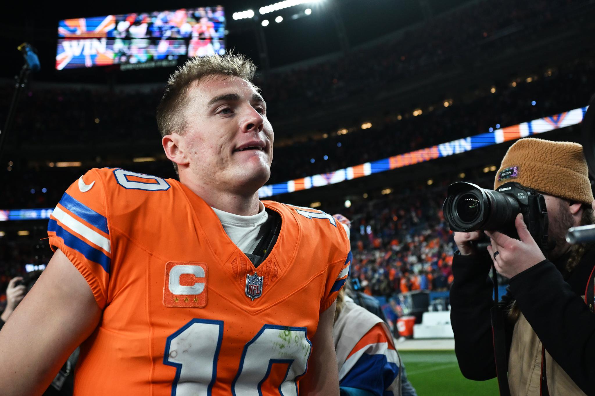 Bo Nix in an orange jersey on the field with a photographer nearby.