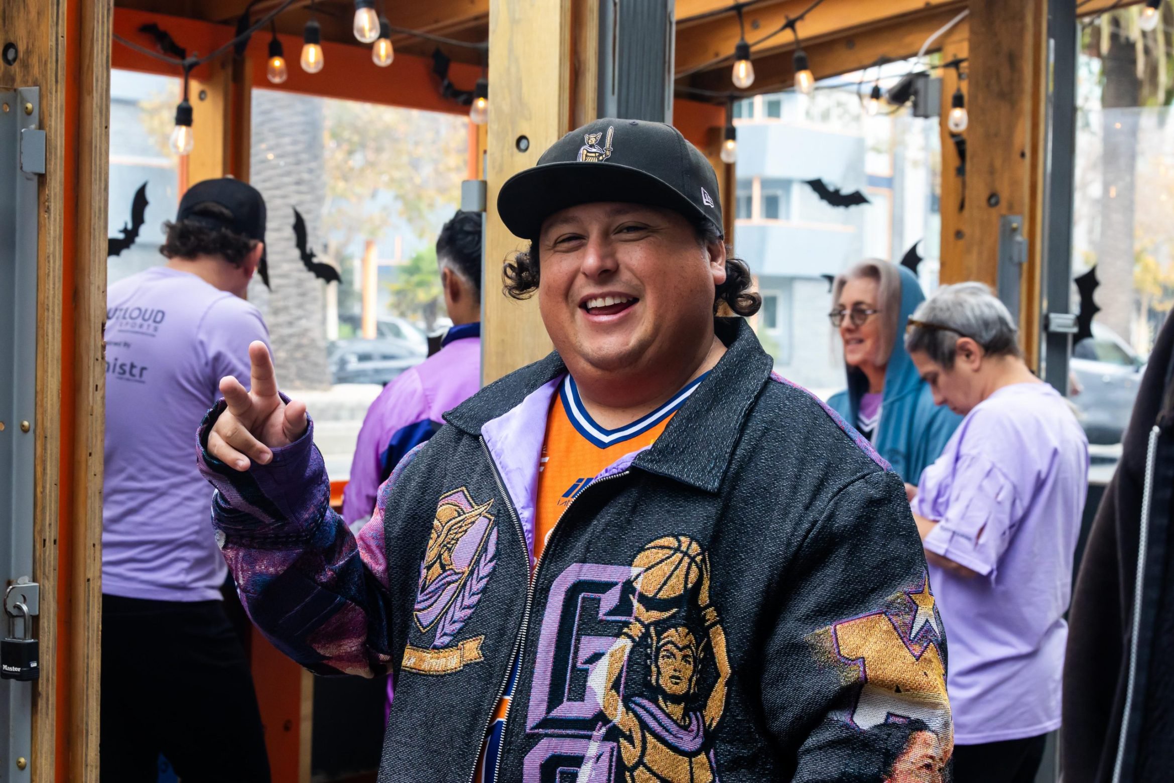 A smiling man wearing a black baseball cap and a patterned jacket makes a peace sign, with blurred people standing in the background.