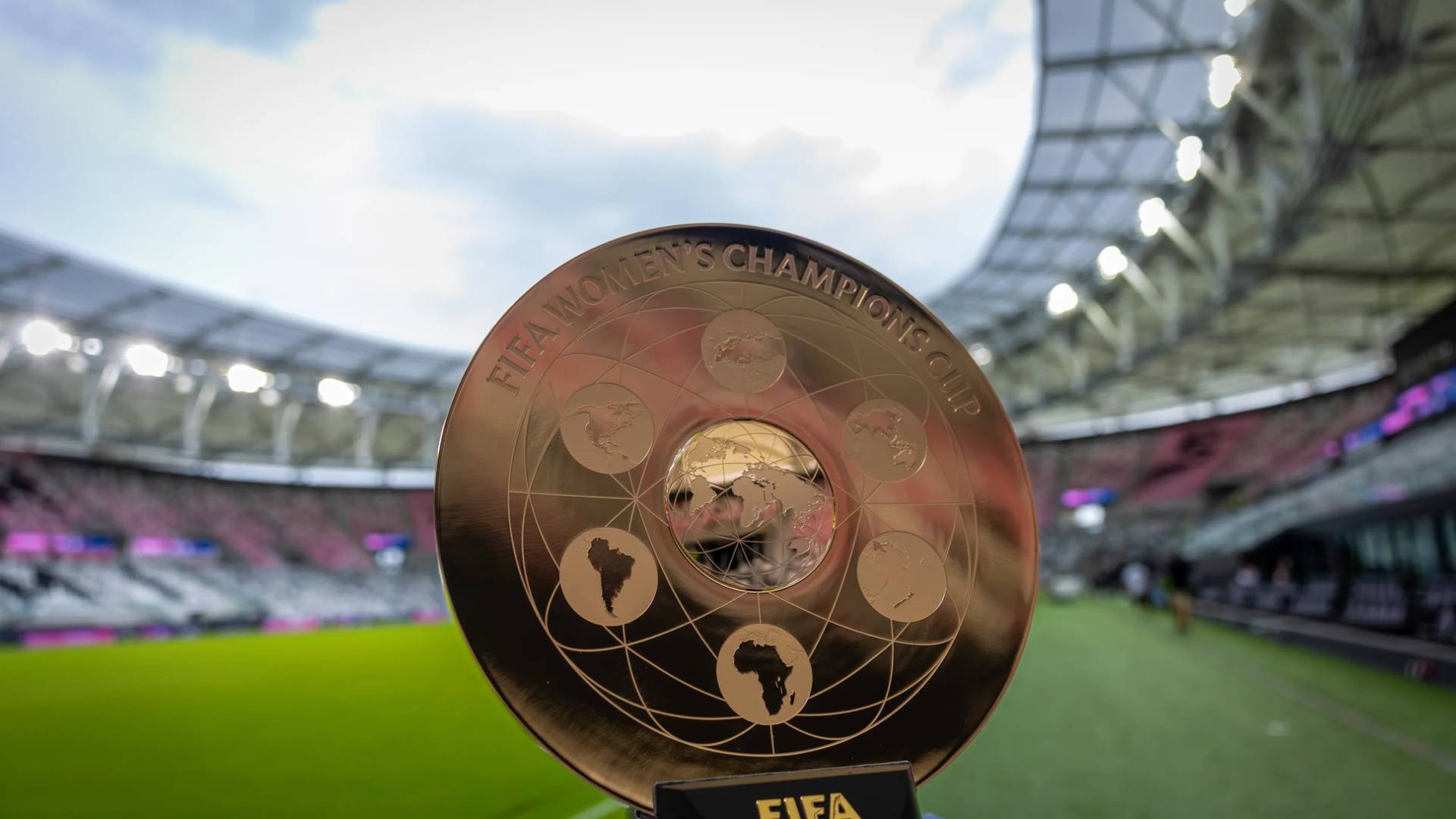 Troféu dourado da FIFA Womens Champions Cup em primeiro plano, com o mapa-múndi e continentes gravados. Ao fundo, um estádio de futebol com gramado verde e arquibancadas vazias, sob um céu nublado