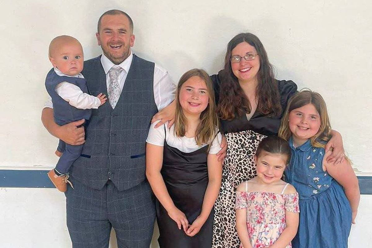 A family of five individuals, including two young girls, a baby, and two adults, are posing together for a photograph in a room with a white wall.