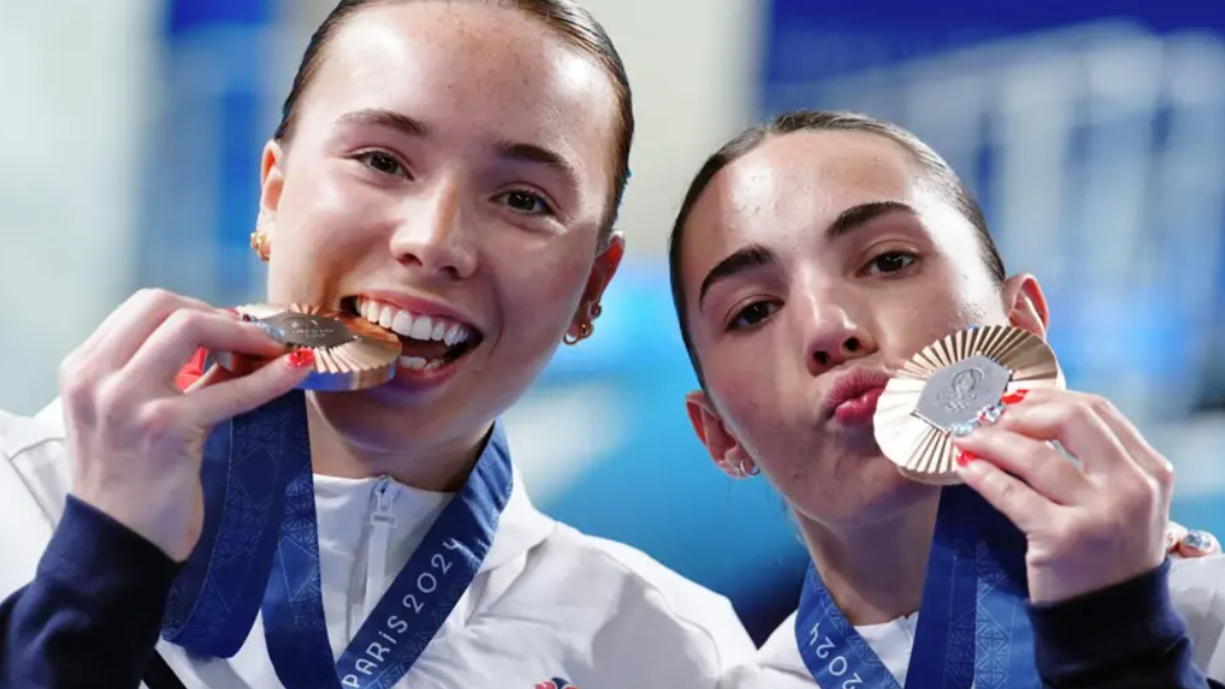 Two young women in white tracksuits smile as they hold up bronze medals from around their neck