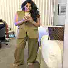 Woman taking a mirror selfie in a bedroom, dressed in a tan pantsuit with loose, sleeveless blazer and wide-leg trousers, holding a phone. Nearby is a designer bag