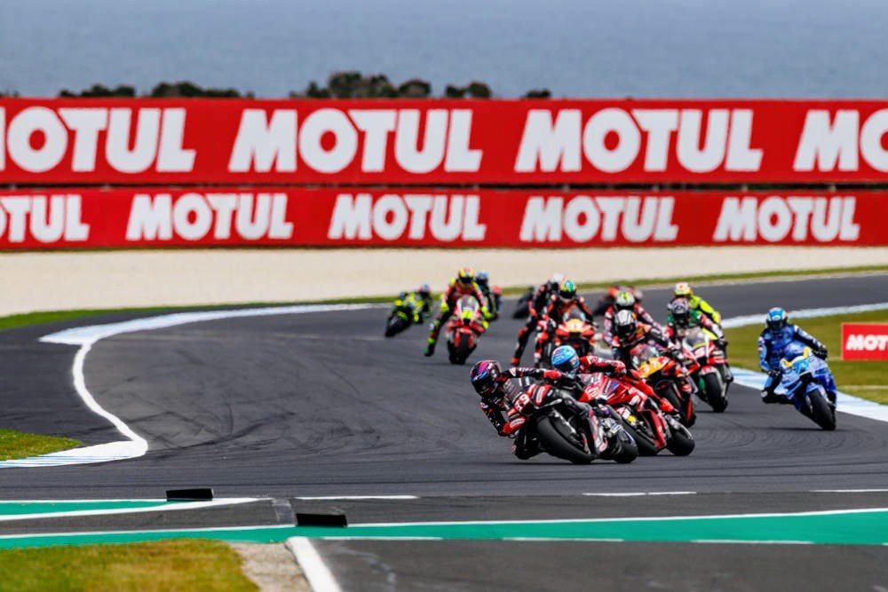 MotoGP riders racing through a corner at the Australian GP, with a red Ducati and a rider in a blue suit on a Suzuki-style bike leading a pack past MOTUL ban…