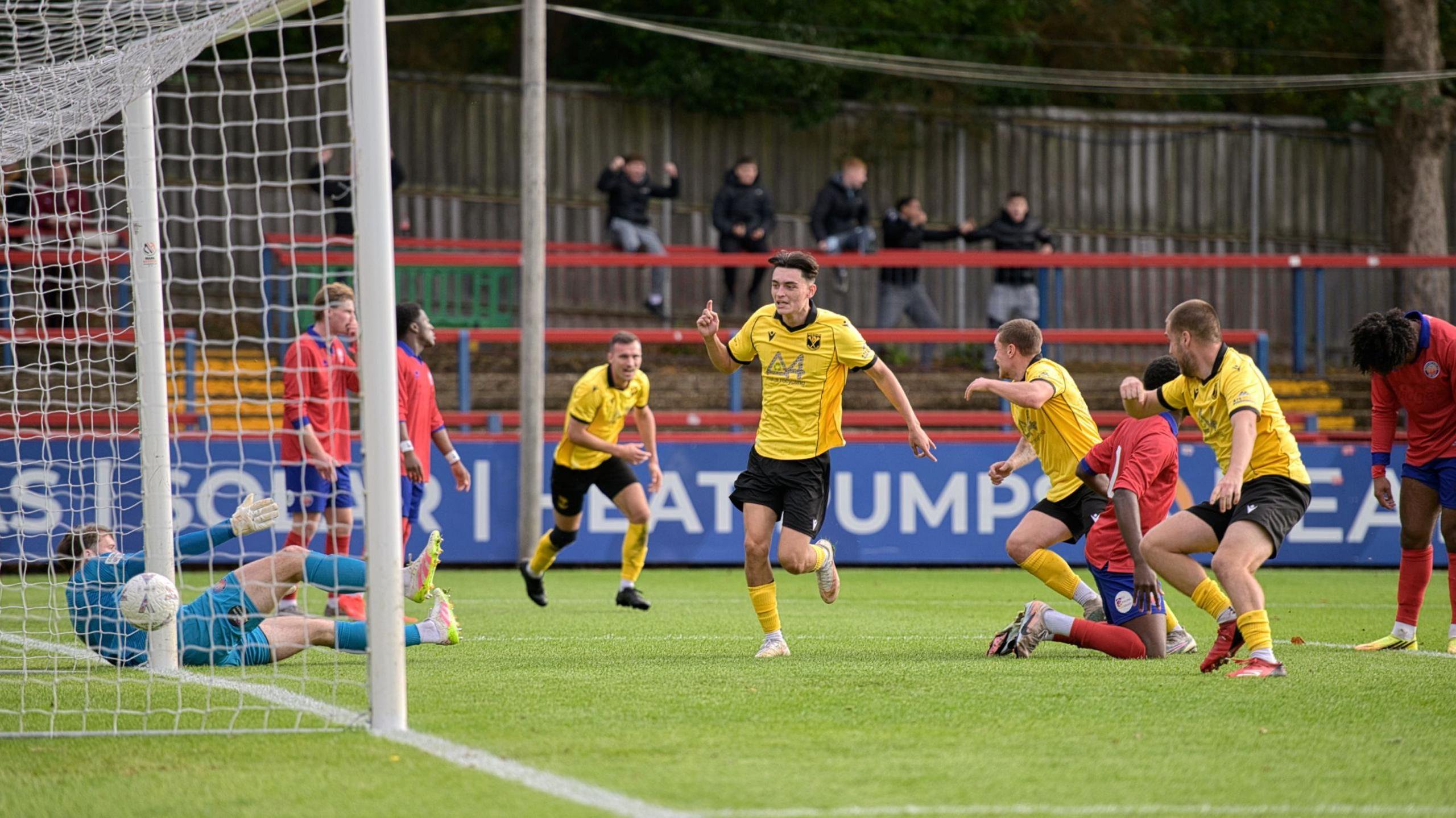 Football players in yellow kit celebrate as they score a goal. Players in red kit hang their heads in defeat.