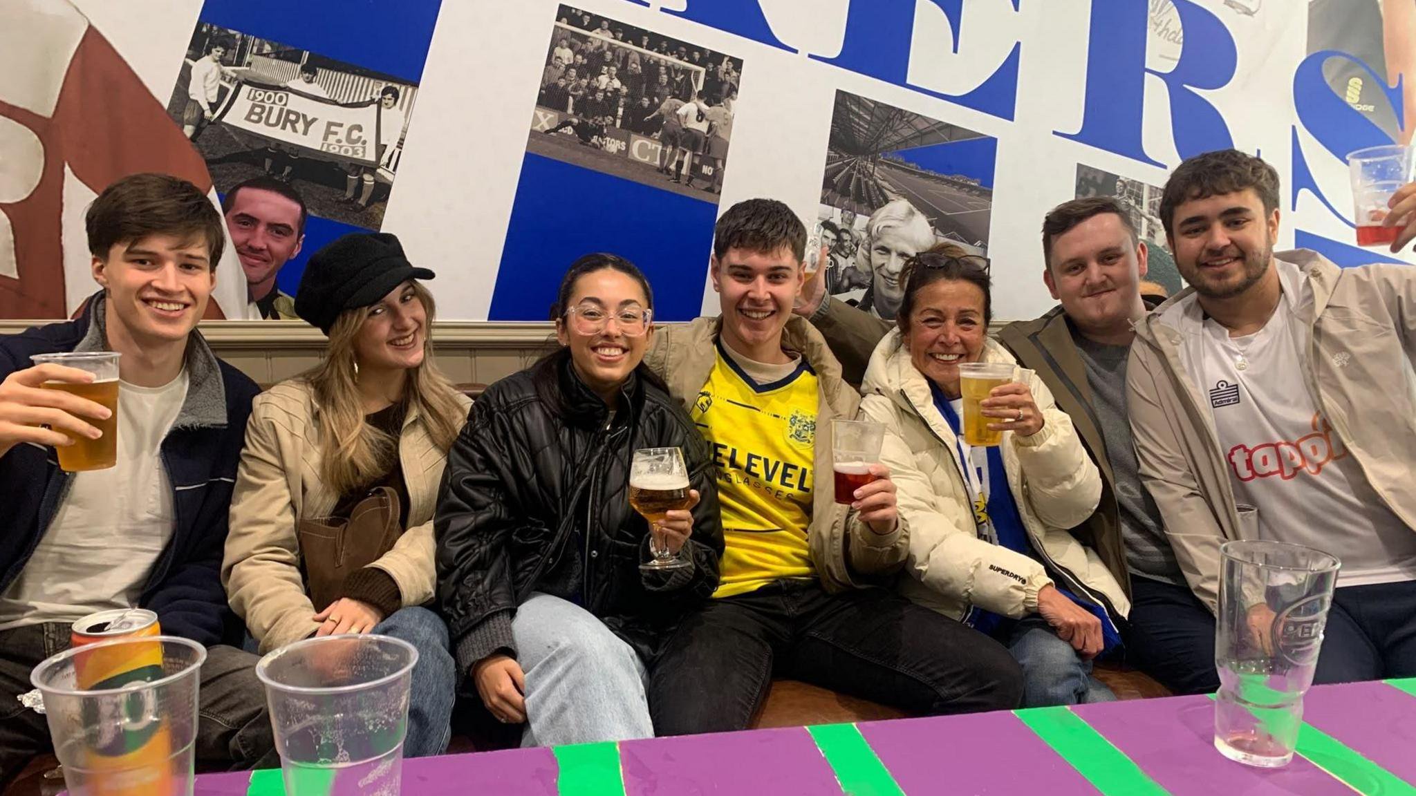 A group of Bury FC supporters sit together inside a bar area, holding drinks in front of a wall featuring historic photographs and large Bury FC-themed artwork.