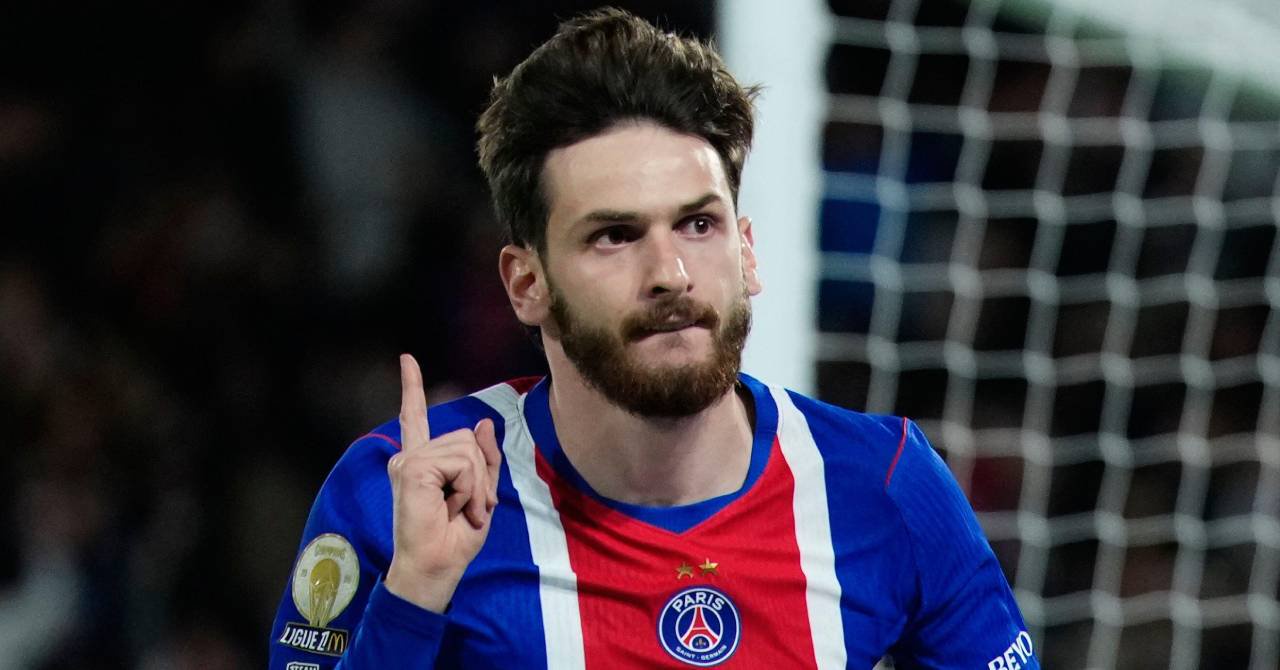 Georgia footballer Khvicha Kvaratskhelia raising his right hand and digit finger to celebrate a goal while playing for Ligue 1 club Paris Saint Germain Football Club while wearing a blue, red and white PSG home shirt in a stadium in front of a white goal post and net. The Ligue 1 logo is visible on his right arm and the club badge is on the chest with a graphic of the Eiffel Tower in France.