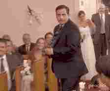 Steve Carell in a suit and tie humorously dances down the aisle at a wedding ceremony with guests and a bride and groom in the background. &quot;Peacock&quot; logo in corner