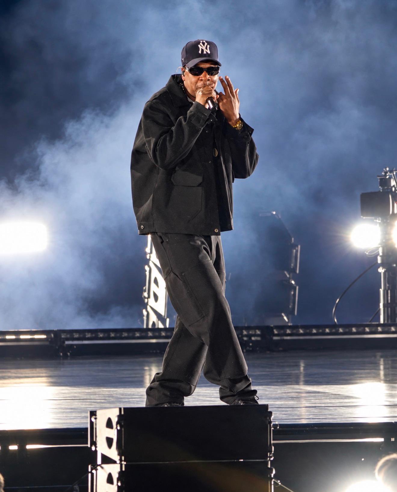 Jay-Z performing on stage during his tour in Los Angeles, wearing a black jacket, sunglasses, and a Yankees cap, surrounded by smoke and stage lights.