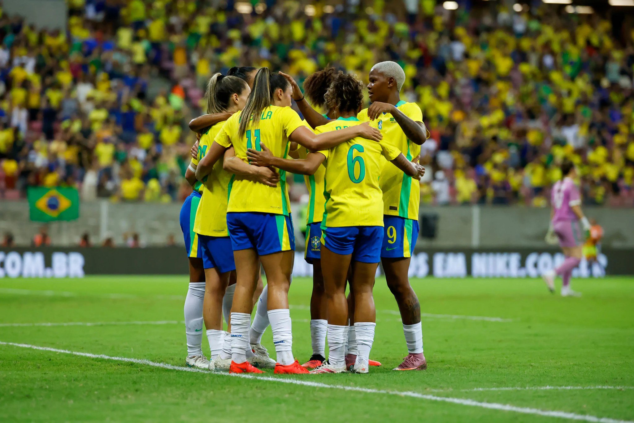 Seis jogadoras de futebol brasileiras, vestindo uniformes amarelos e shorts azuis, se abraçam em círculo no gramado verde de um estádio, celebrando. A torcida, em sua maioria com roupas amarelas, aparece desfocada ao fundo, assim como uma bandeira do Brasil e uma goleira de uniforme roxo.