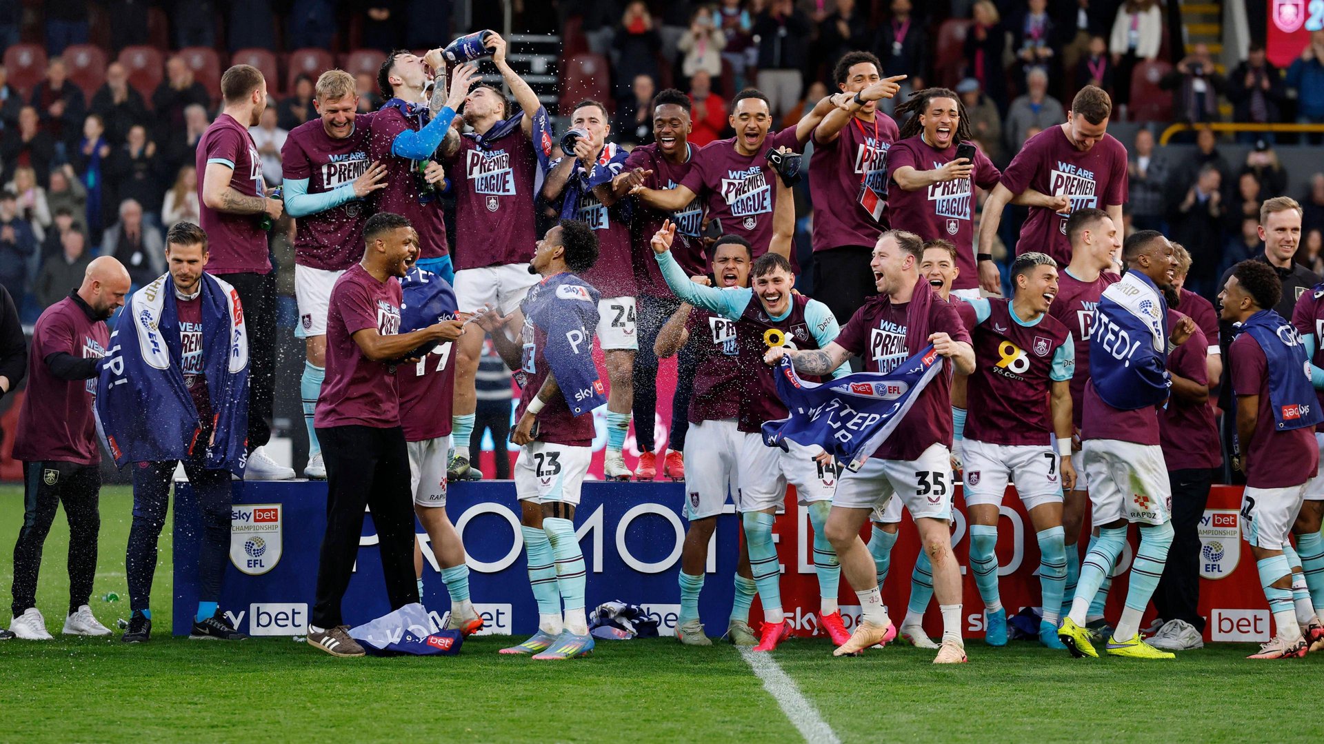 Burnley players celebrate winning promotion to the Premier League in 2025 by swigging from bottles of champagne and wearing claret t-shirts with Premier League written on the them