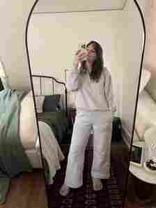 Person wearing a light gray tracksuit takes a mirror selfie in a bedroom. The room features a bed with green bedding, a nightstand, and a white sofa.