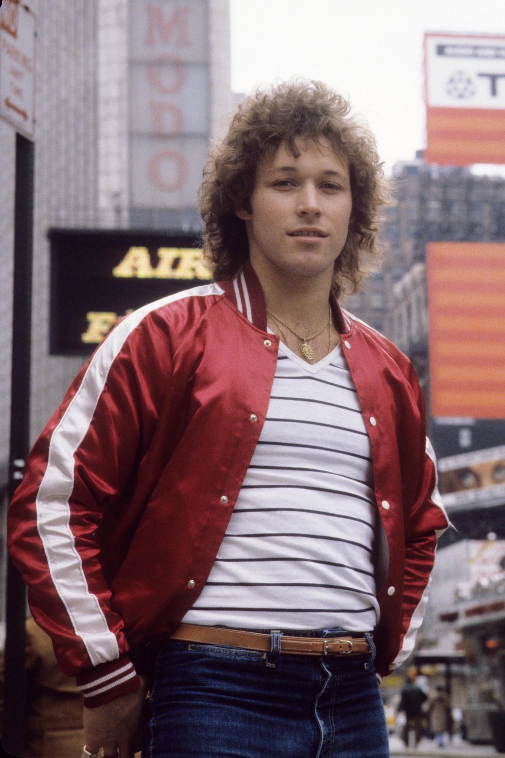 Canadian hockey player Ron Duguay poses for a portrait on a New York City street, wearing a red satin jacket, striped shirt, and jeans.