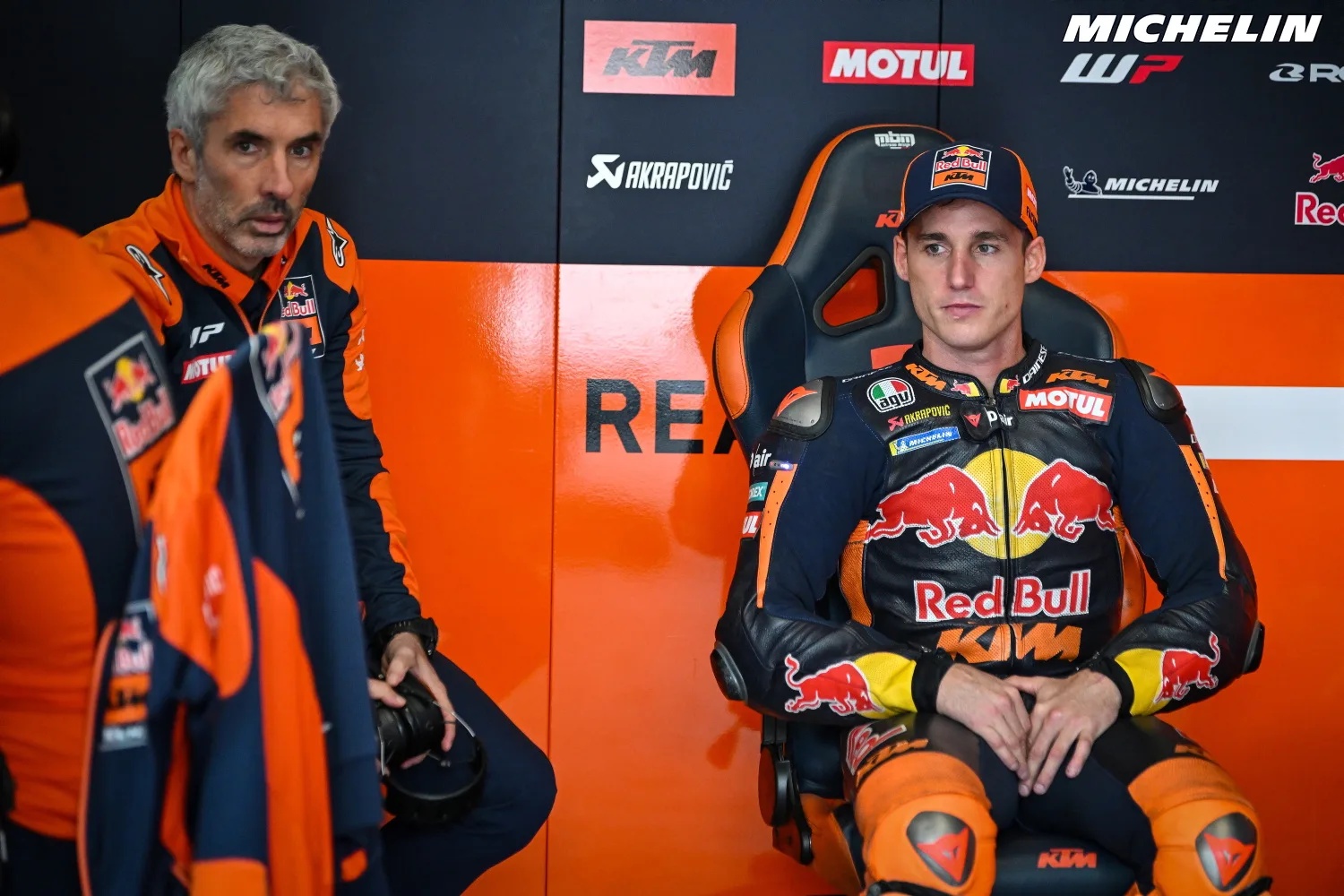A Red Bull KTM MotoGP rider in orange and black leathers sits in the team garage, with a team member standing beside him.