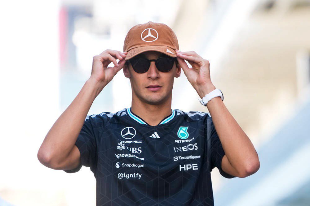 A man in a Mercedes-AMG Petronas F1 team shirt and brown Mercedes cap adjusts his cap and sunglasses in the paddock