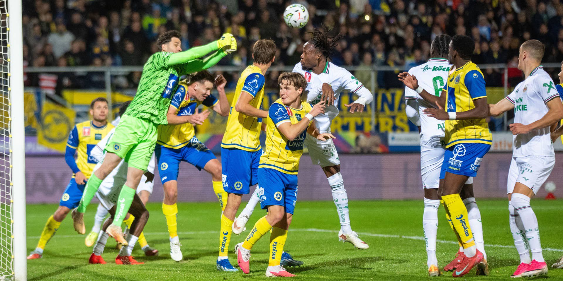 Fußballspiel: Torwart springt im Strafraum, mehrere Spieler kämpfen um den Ball.