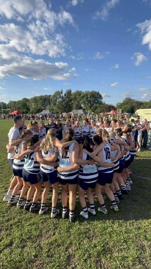 A group of players in white-and-navy striped sports uniforms stand in a tight huddle on a grass field, arms around each other, during an outdoor sporting event. Spectators, other participants and tents are visible in the background, with trees beneath a blue sky dotted with clouds.