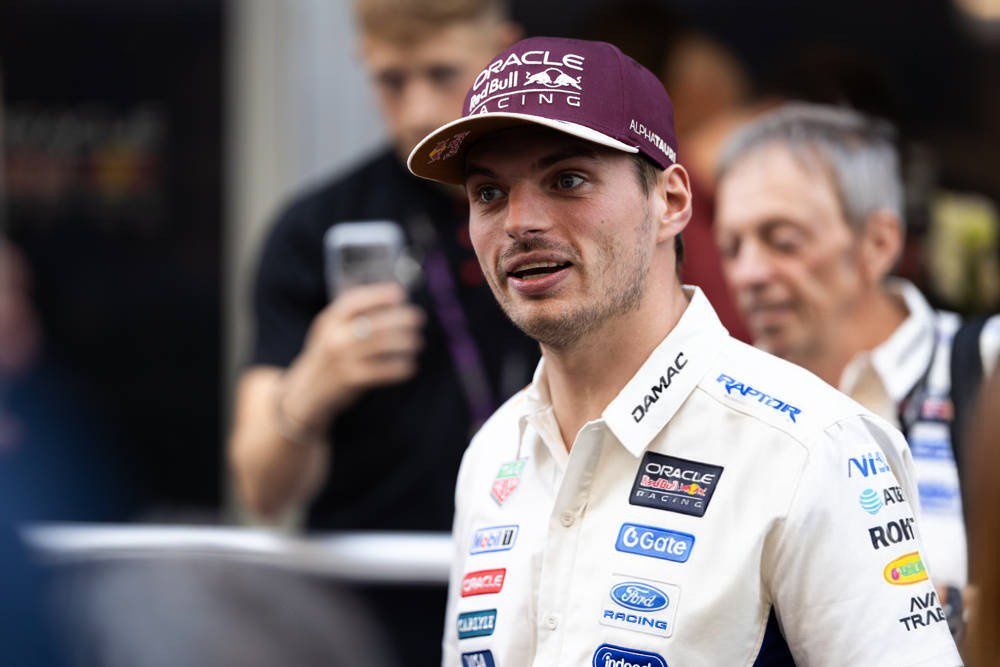Driver in a white Oracle Red Bull Racing shirt and maroon cap with sponsor logos, speaking in the Formula 1 paddock, with people behind him.