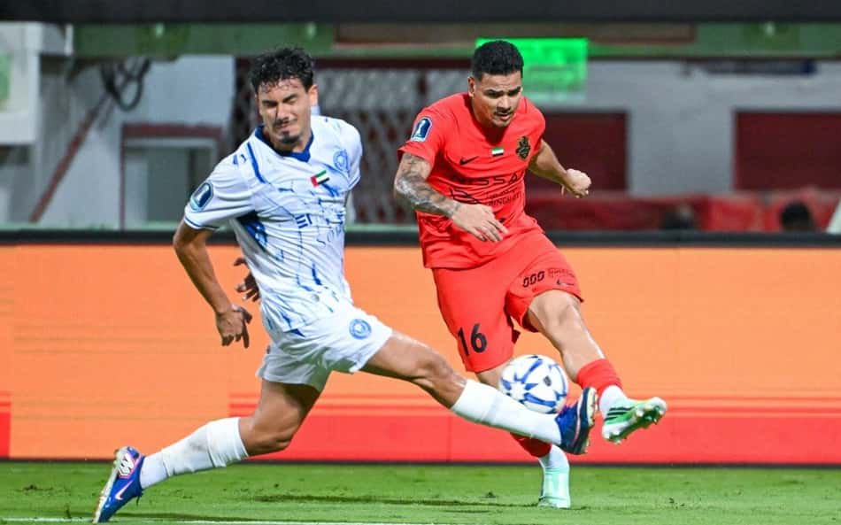 Dois jogadores disputam a bola em campo; um veste uniforme laranja com o número 16, o outro branco com detalhes azuis.