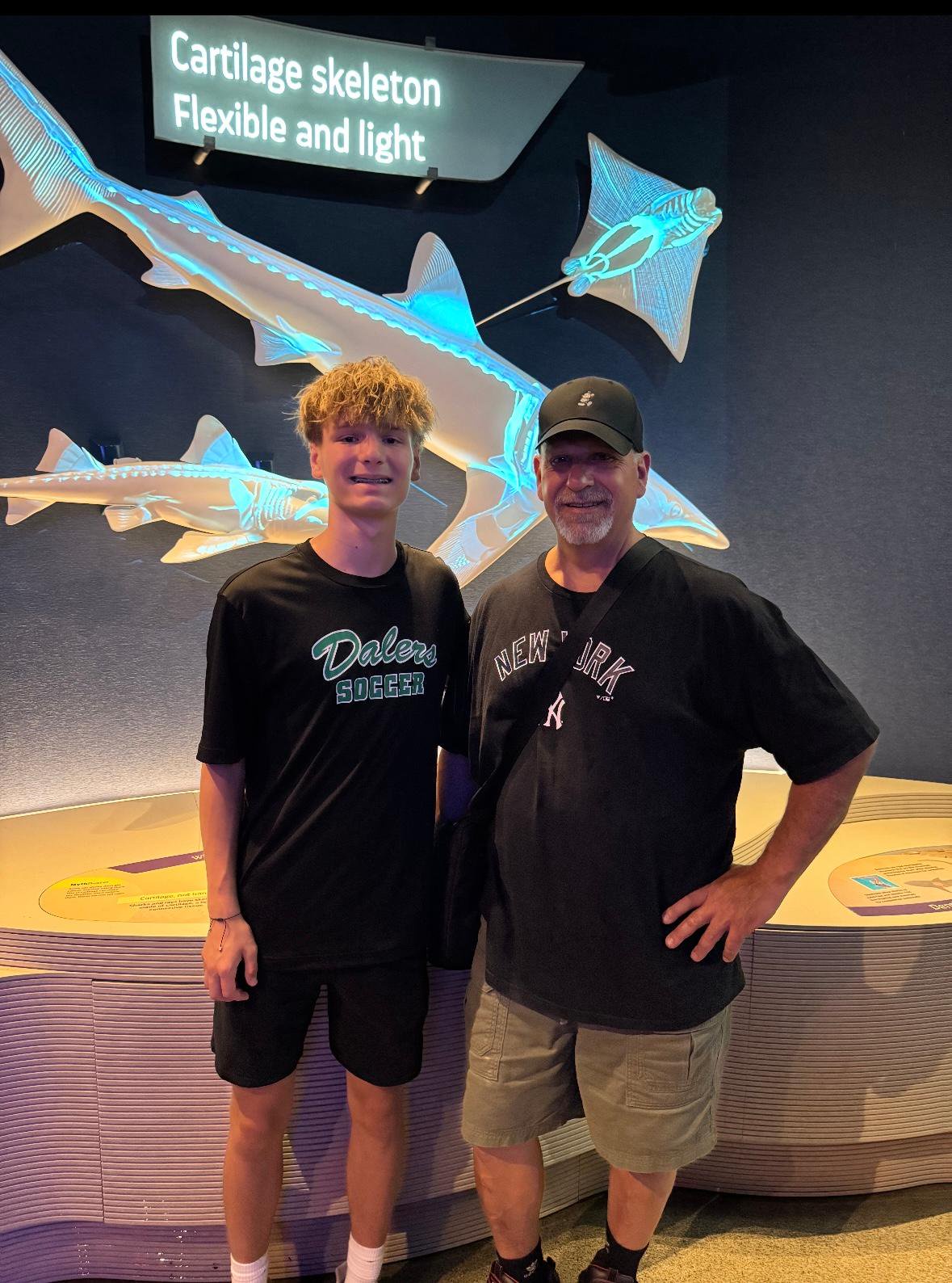 A boy and an older man stand smiling in front of a museum exhibit about cartilaginous skeletons, with glowing models of fish skeletons in the background.
