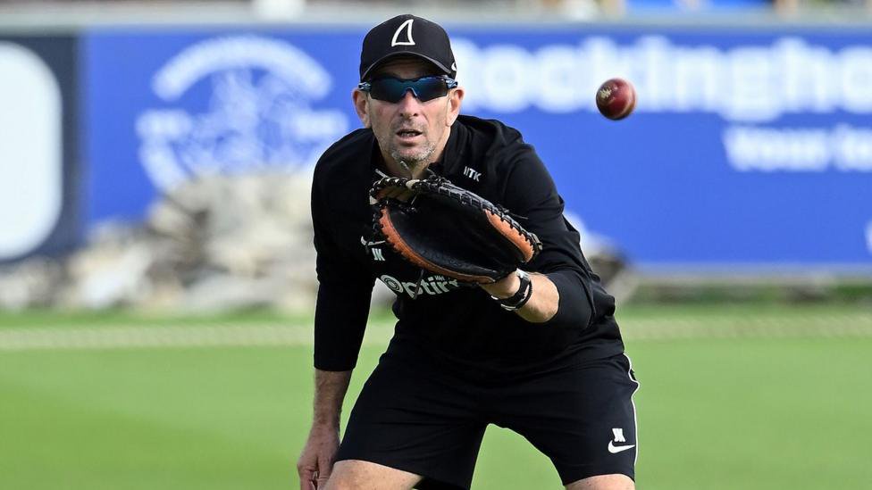 James Kirtley in dark Sussex kit and cap, with shades, prepares to catch a cricket ball in a mitt