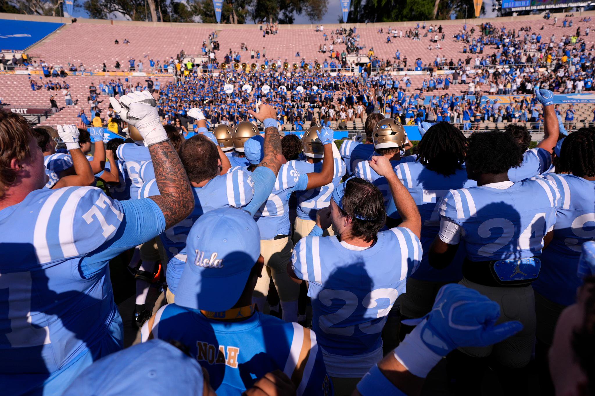 UCLA football players raise their arms in victory facing the marching band and crowd in the stadium.