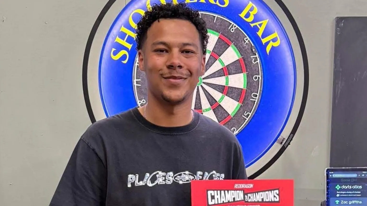 Zac Griffiths Zac Griffiths stands in front of a dartboard that reads "Shooters bar". He has dark brown hair and is smiling. He holds a red sign reading "Champion of Champions".