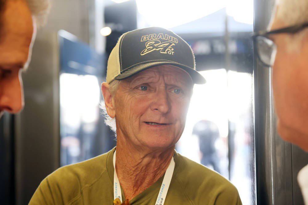 Kevin Schwantz, in a black cap with "Brand 34 GT" logo and olive shirt, speaking with a man in the paddock.