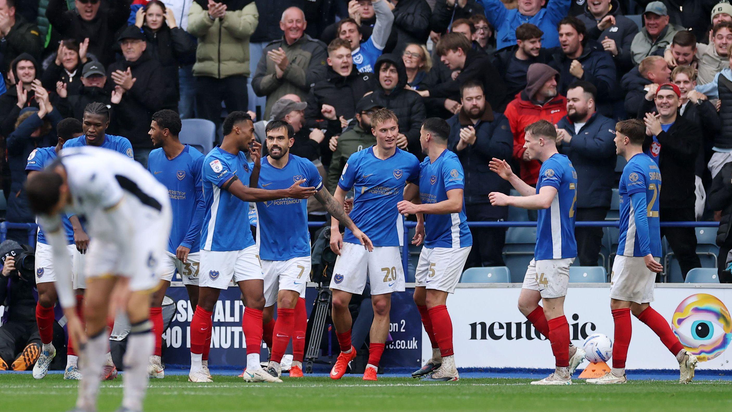 Portsmouth players, in their blue, white and red home kit, celebrate the winning goal against Millwall. The Pompey fans can be seen celebrating in the stands behind them.
