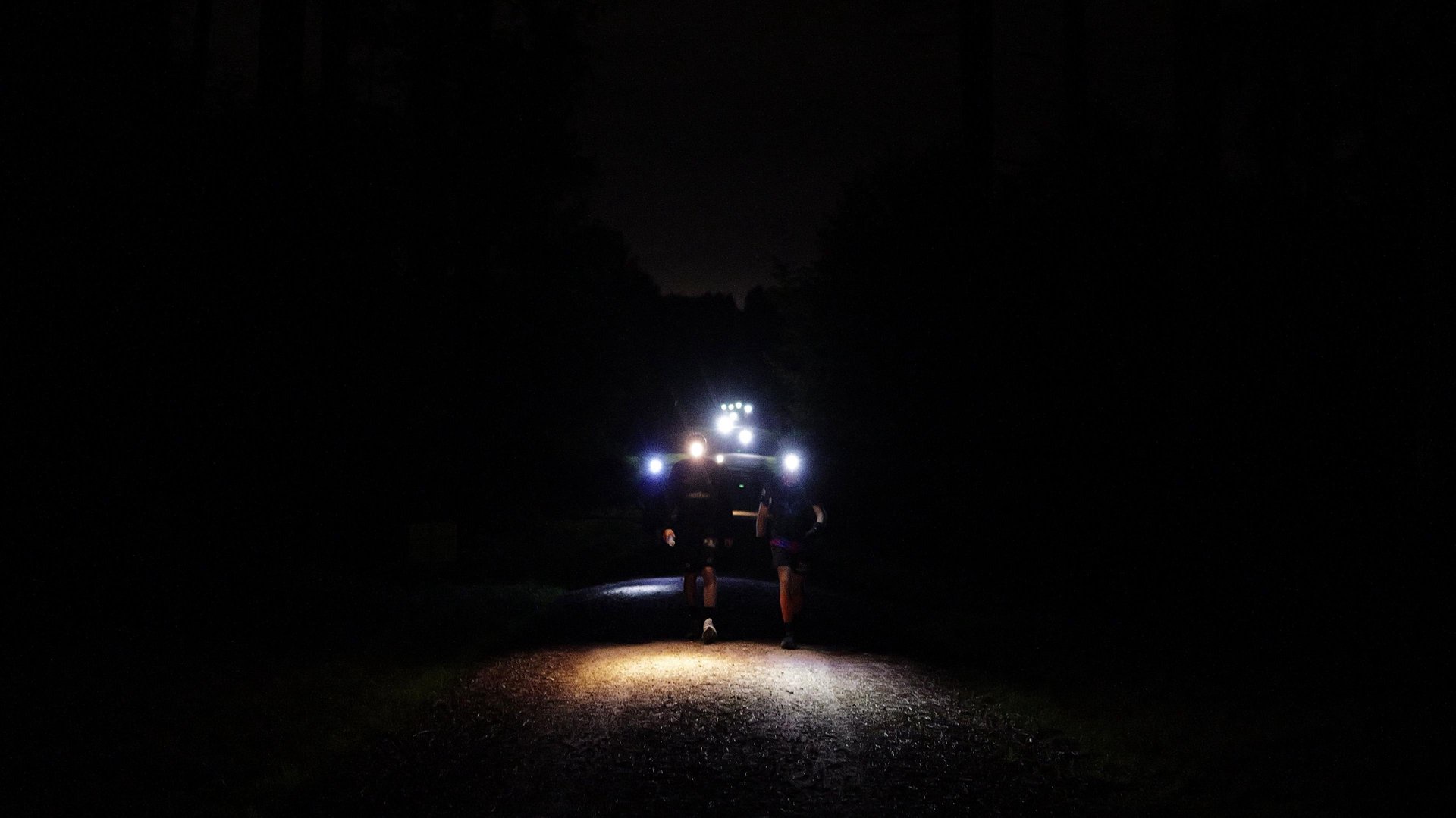 It is night time and is pitch black. There are two runners running side by side and they both have head torches on.
