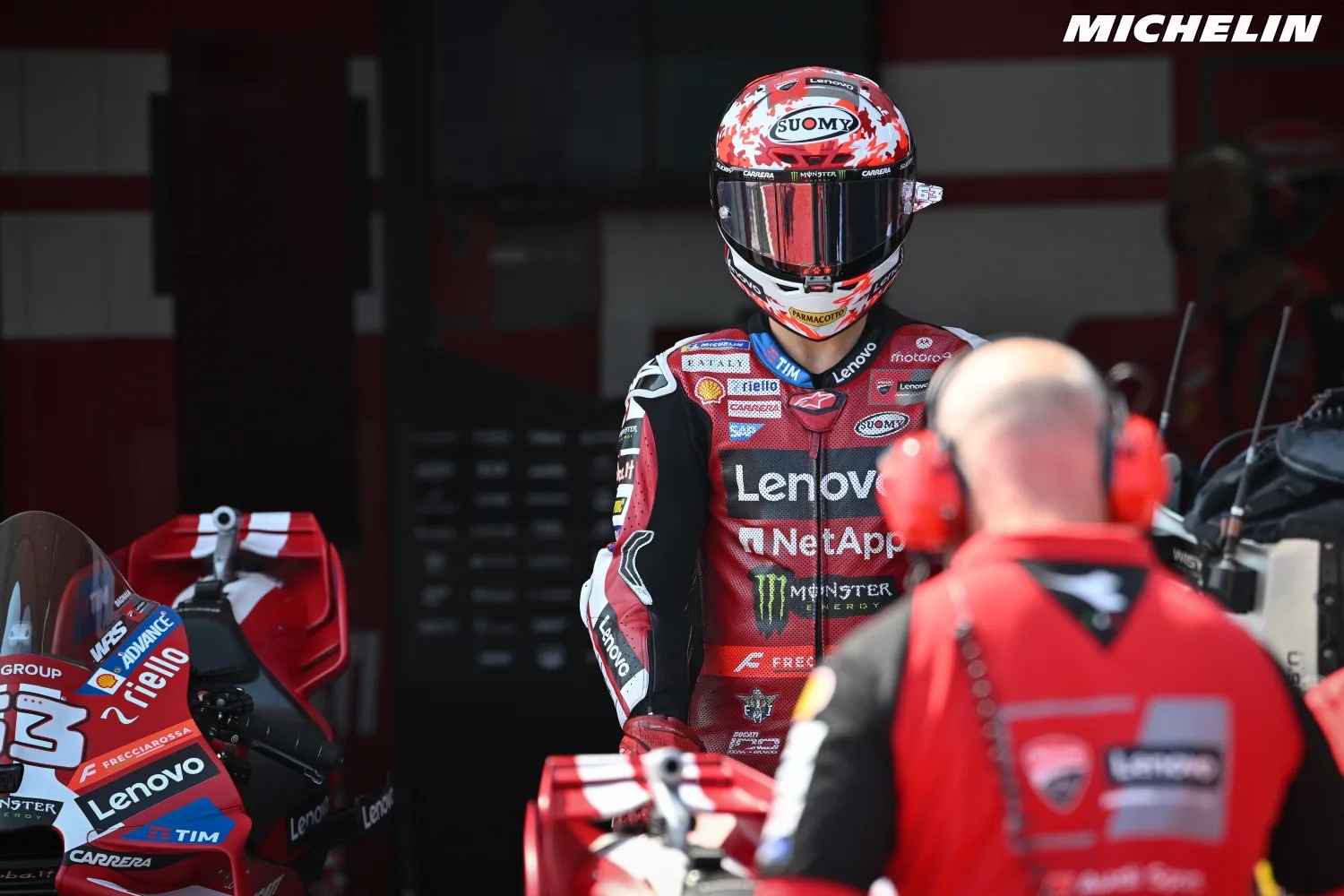 Ducati rider in red Lenovo leathers and Suomy helmet in the MotoGP pit garage, with a crew member in red in the foreground