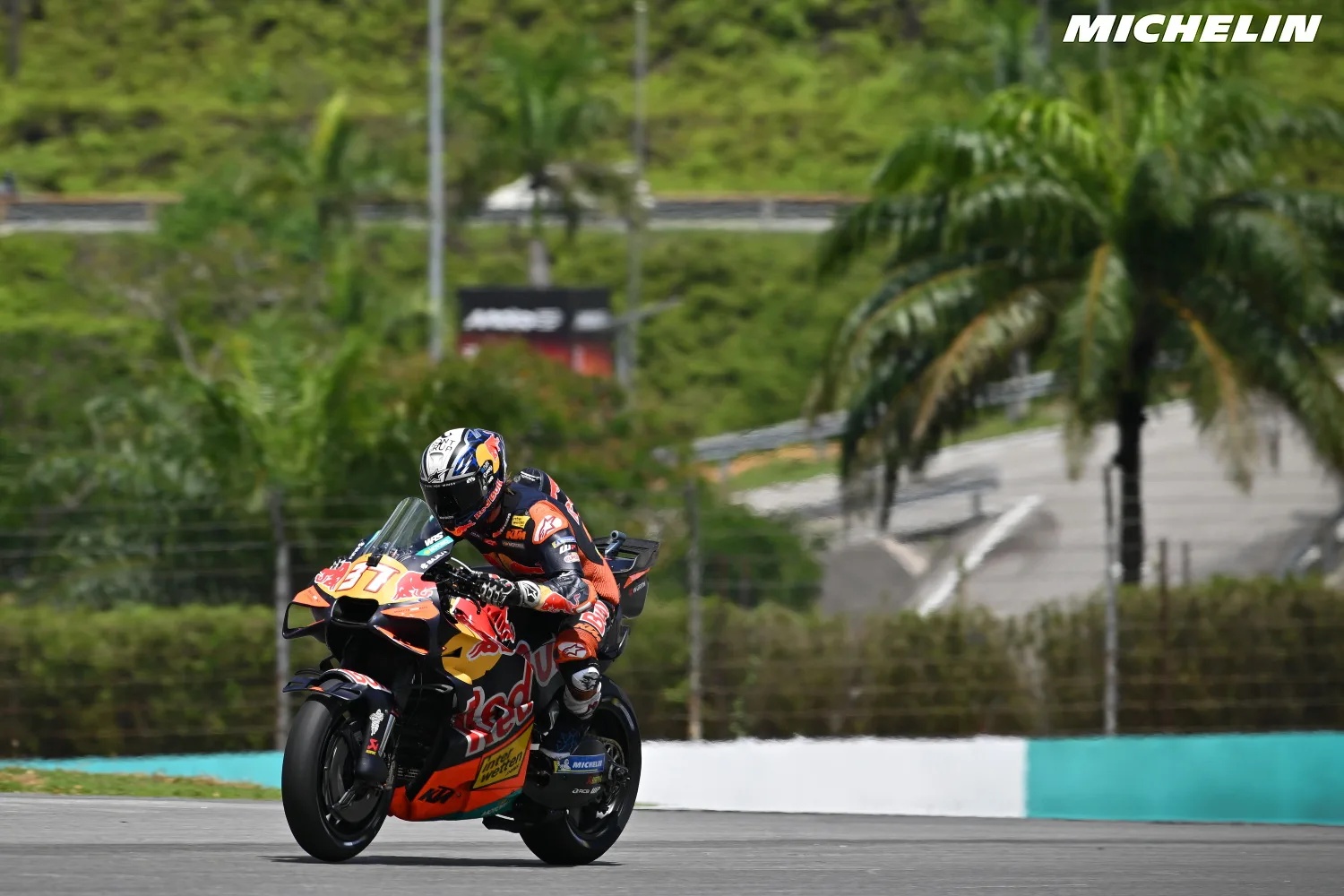 A rider on a Red Bull KTM MotoGP bike with orange-and-black livery and a number 37 fairing, leaning through a corner at a tropical circuit.