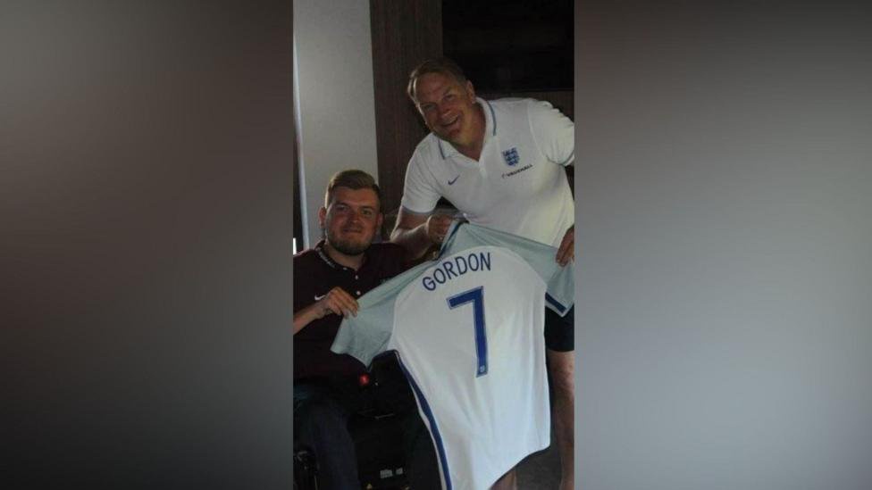 Chris holds up an England shirt with the name Gordon on the back with his father also holding it. Colin is wearing an England top and leans in to his son, in a wheelchair, for the photo
