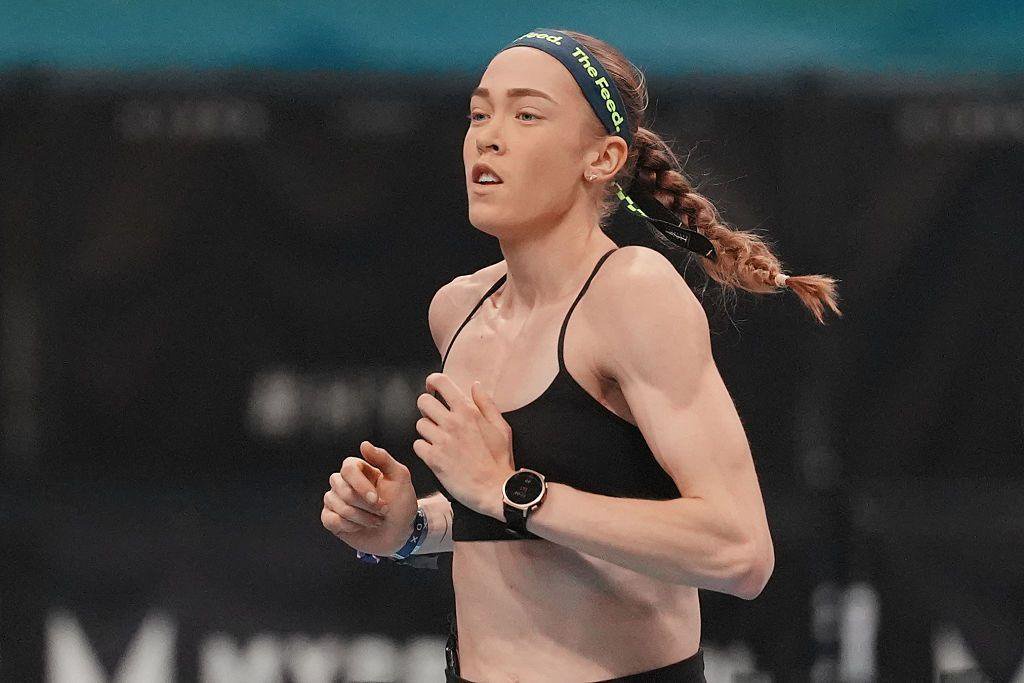 Joanna Wietrzyk wears a black sports bra and a dark headband with the words 'the feed' printed in green letters. Her hair is tied back into a plait and she is mid-run.