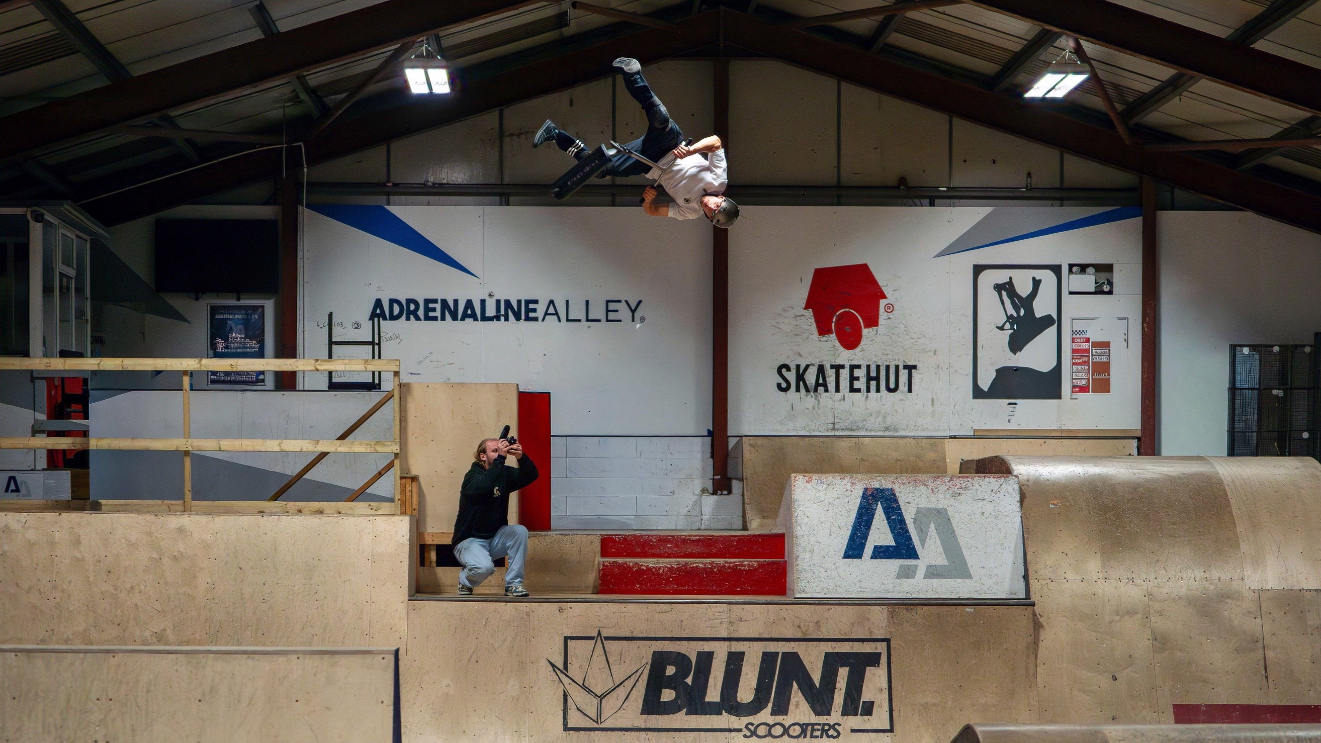 A man taking part in a scooter jump in mid air, holding on to a scooter. He is upside down and wearing a white T-shirt and black trousers.