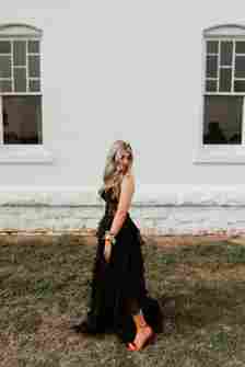 A woman with long, wavy hair stands on grassy ground in front of a white building with two windows. She is wearing a black, layered strapless gown with red high heels and a wrist corsage. She is looking down while slightly turning away from the camera.