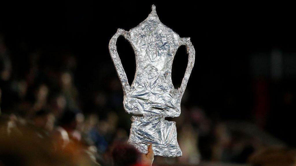 A cardboard cut-out of the FA Cup, wrapped in tinfoil. It is held up by a just-glimpsed hand.