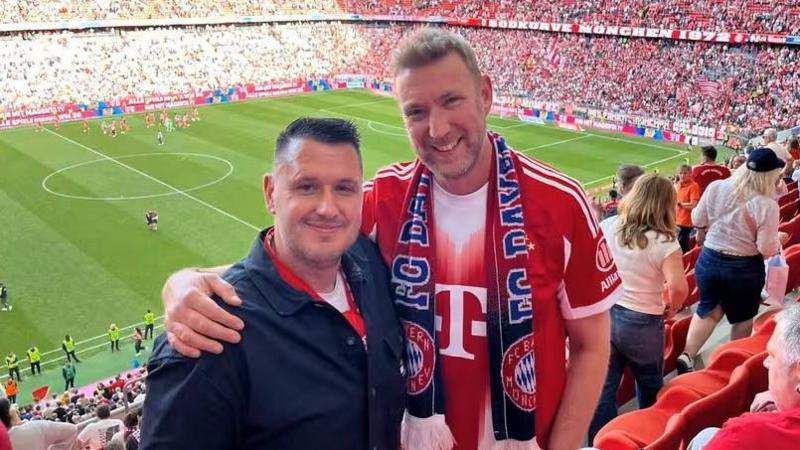 A man with black hair and a black shirt stands next to a taller man who has his arm around his shoulder. The man on the right is wearing a red shirt and a Bayern Munich scarf.  They are standing in a football stadium. 