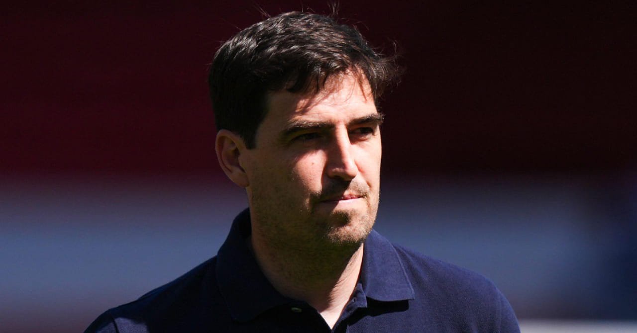 Football manager Andoni Iraola looking to his left while pursing his lips in an outdoor setting with sunshine across his face. The coach is wearing a dark polo shirt