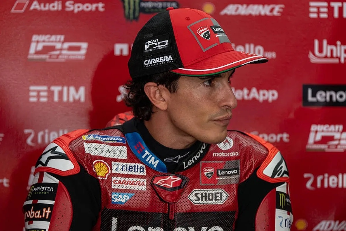 A Ducati rider in a red and black Ducati Lenovo leather suit and cap in the MotoGP paddock, with team sponsor backdrop behind him