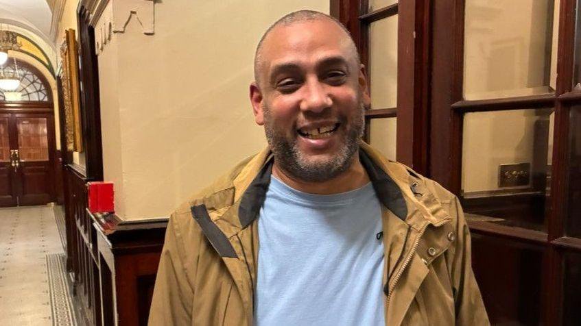 Darryl Shellard has short grey and black hair and a beard. He is wearing a light  blue t-shirt and beige jackets and is stood inside Liverpool Town Hall next to a glazed wooden door.