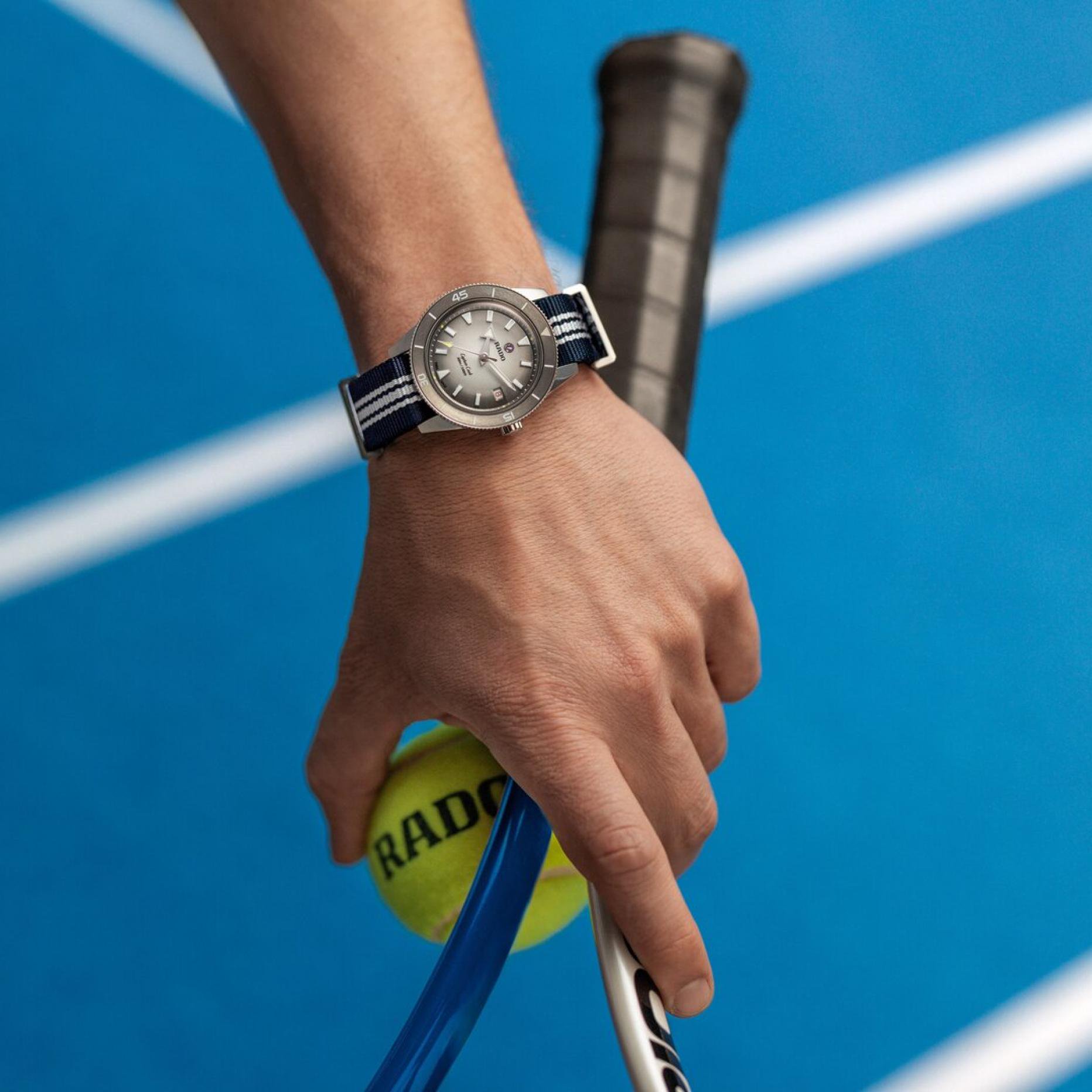 Eine Hand trägt einer Rado Armbanduhr mit blau-weißem Textilarmband, sie hält einen Tennisschläger sowie einen gelben Tennisball.