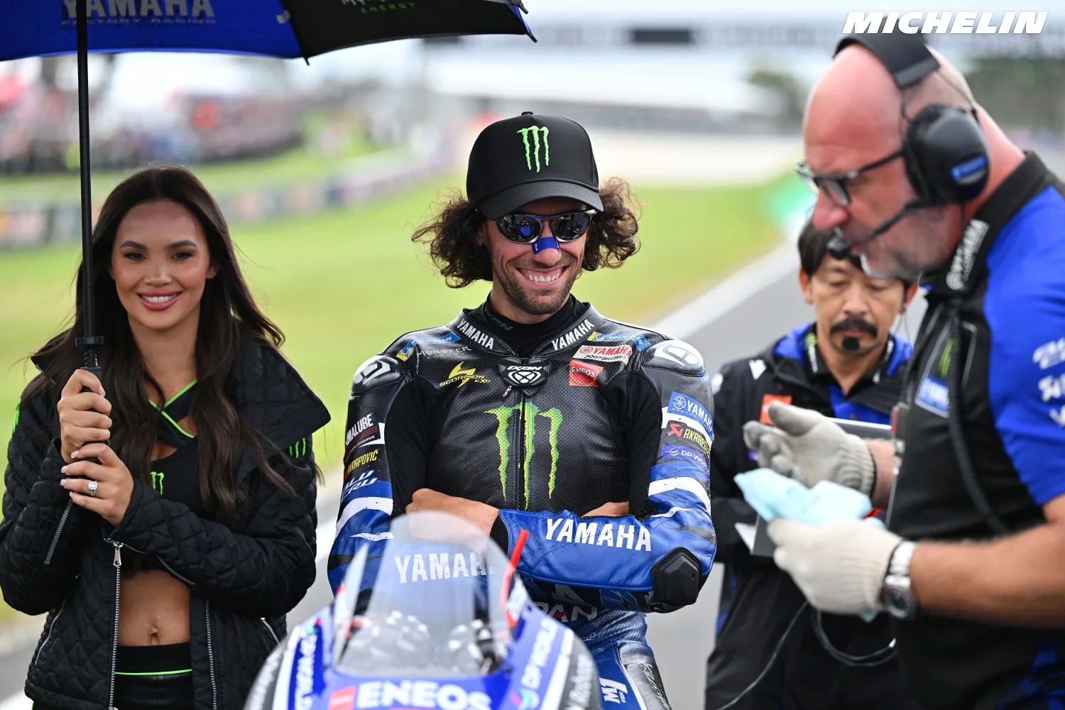 Rider in black Yamaha leathers and Monster cap stands on the MotoGP grid beside a Yamaha umbrella-holder and crew member with headset.