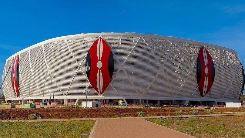 A wide shot of the outside of a football stadium showing three giant Kenyan shields in red, white and black on top of a white wraparound exterior. A clear blue sky is visible above the stadium while partially finished groundsm with scrubby grass, are being worked on in the foreground
