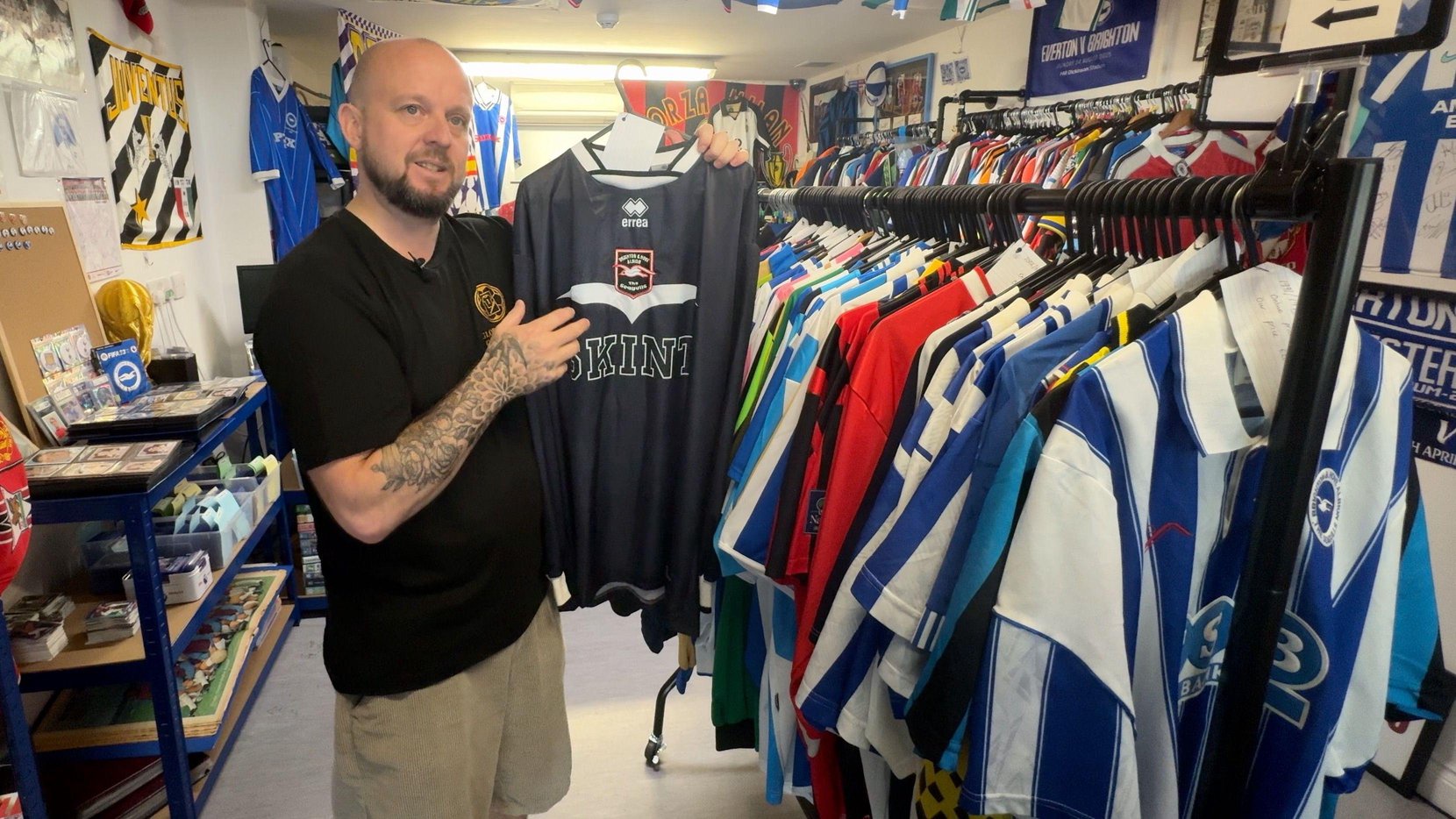 A bald man wearing a black t-shirt, with tattoos on his arm, holding up a black football shirt in his shop. There is a rail of football shirts to the right, with other football memorabilia in the background