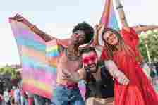 A group of three people celebrates Pride, waving a rainbow flag. One person wears heart-shaped glasses, another has dreadlocks and a red dress, and the third is smiling broadly