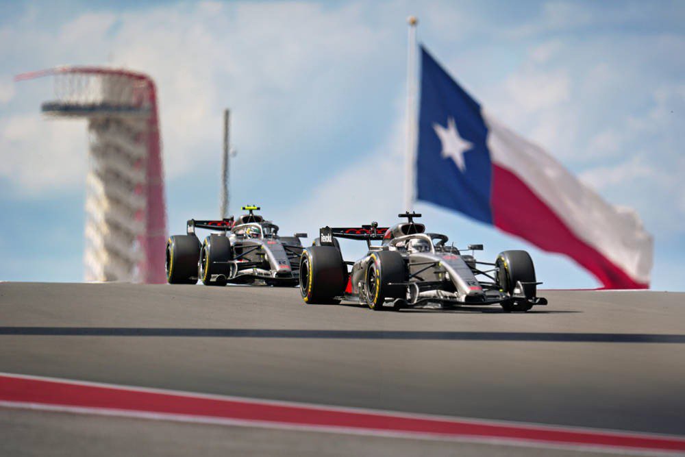 Two Audi F1 cars racing on the Circuit of the Americas track in Texas, with a Texas flag and the track's tower behind them.