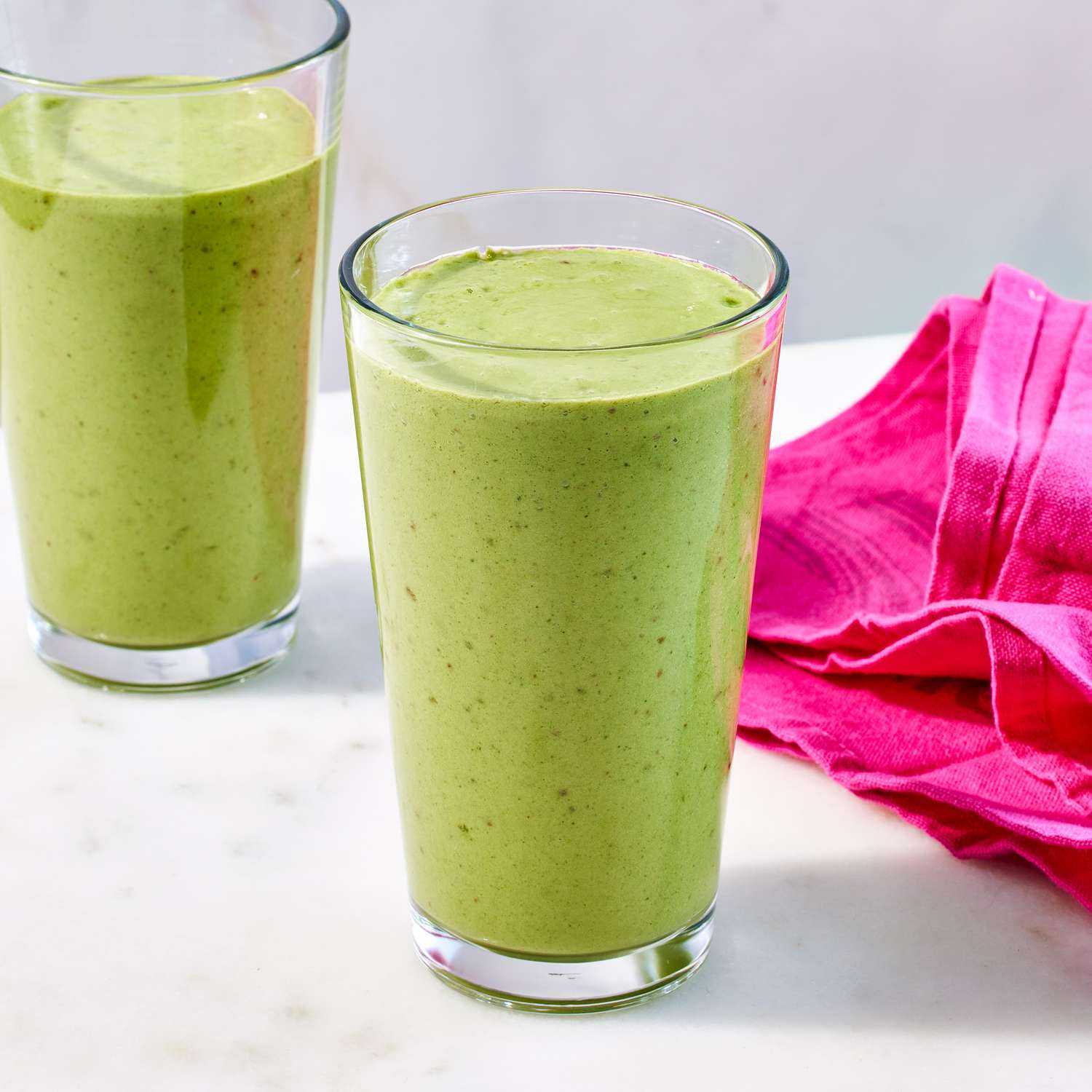 two tall pint glasses filled with green smoothie next to hot pink napkin