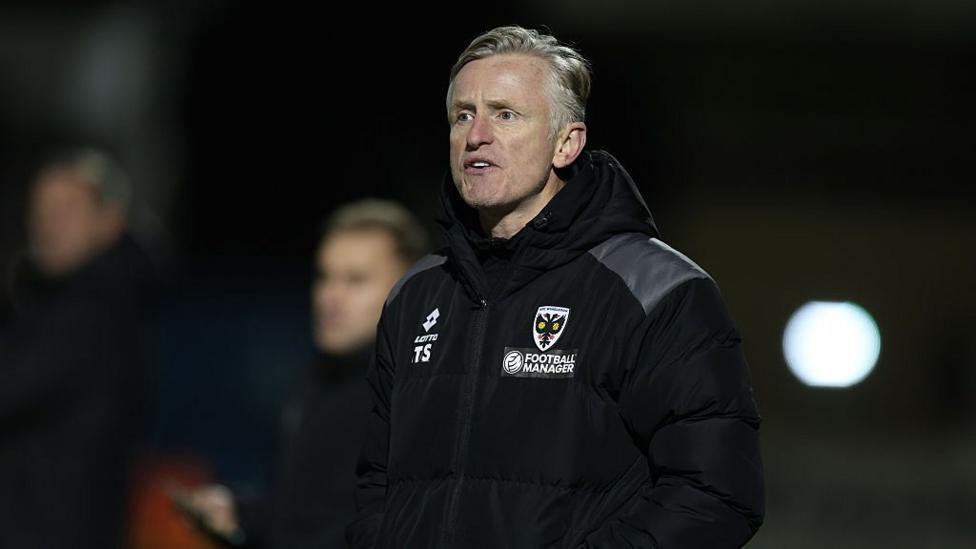 AFC Wimbledon assistant manager Terry Skiverton looks on from the touchline wearing a black coat